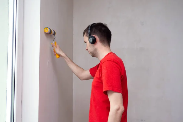 Homme amorçant le mur avec un rouleau et écoutant de la musique dans le casque. Réparation de l'intérieur. Jeune décorateur masculin peignant un mur dans son appartement. — Photo