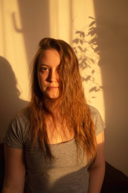 Gün batımının sıcacık ışıklarıyla kameraya bakan genç bir kadın. İçerideki kızın sıcak sakin portresi. Yüzünde ışık ve gölge.