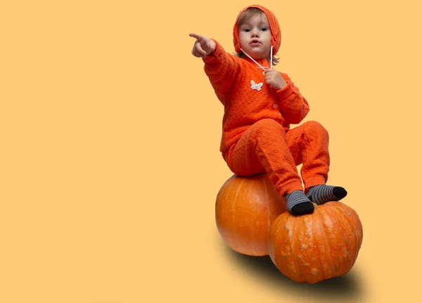 Kid girl in an orange suit sits on pumpkins and points a finger copy space. place for text halloween. — Stock Photo, Image