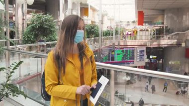 Havaalanında ya da tren istasyonunda yüzü tıbbi maske takan sarı elbiseli, modern turist kadın. Koronavirüs salgını ve karantina kovid 19 'un bitiminden sonraki turizm boyunca seyahat edin..