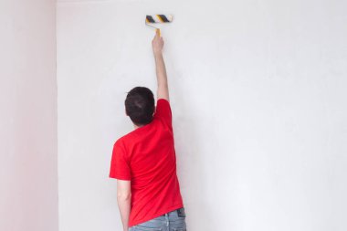 Duvarı silindirle kaplayan bir adam. İçerisinin tamiri. Genç erkek dekoratör boş odada bir duvar boyuyor. Yenileme işini kendin yap..
