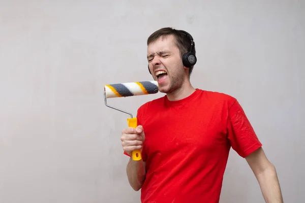 Hombre caucásico feliz cantando canción usando rodillo cepillo como un micrófono. Joven macho divertirse durante la reparación del hogar y la sala de renovación. — Foto de Stock