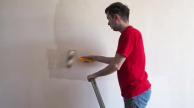 Duvarı silindirle kaplayan bir adam. İçerisinin tamiri. Genç erkek dekoratör boş odada bir duvar boyuyor. Yenileme işini kendin yap.