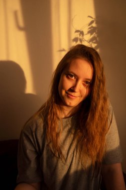 Gün batımının sıcacık ışıklarıyla kameraya bakan genç bir kadın. İçerideki kızın sıcak sakin portresi. Yüzünde ışık ve gölge.