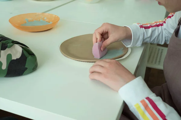 Child painting with sponge on clay pottery plate in ceramic workshop studio. Development of art and painting in children. Close-up — Stock Photo, Image