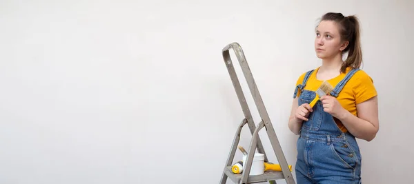 Una mujer pensativa con pincel y escalera se ve soñadoramente en una habitación con una pared sin pintar con espacio para copiar. Independiente hembra sola hace reparaciones de bricolaje en su apartamento — Foto de Stock