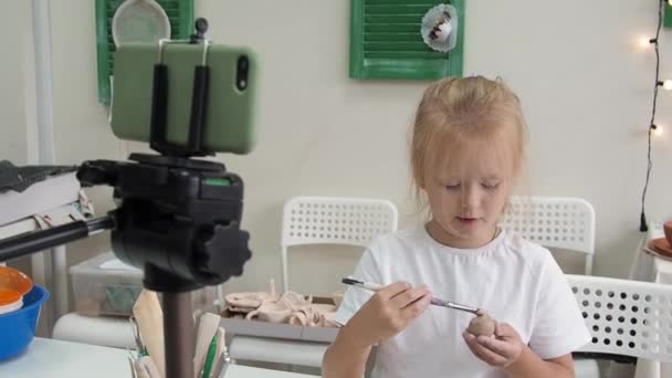 Chica vlogger grabación de vídeo con arcilla de modelado y hablando para la cámara del teléfono inteligente. Blogger infantil en un estudio de cerámica creando contenido interesante para niños — Vídeos de Stock