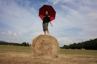 Yazın Toskana 'da saban tarlasında saman topunda şemsiyenin altında güneşin tadını çıkaran genç bir kadın.
