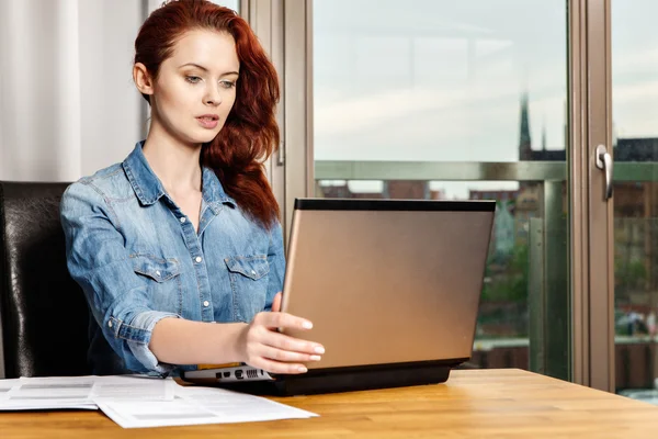 Red-haired young business woman or student girl working with documents and laptop near window — Stock Photo, Image