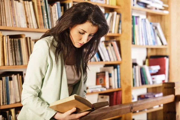 Молода жінка читає книгу перед книжковими полицями — стокове фото