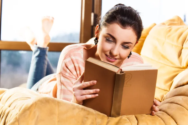 Kobieta leżąc na jej kanapie czytając książkę w domu — Zdjęcie stockowe