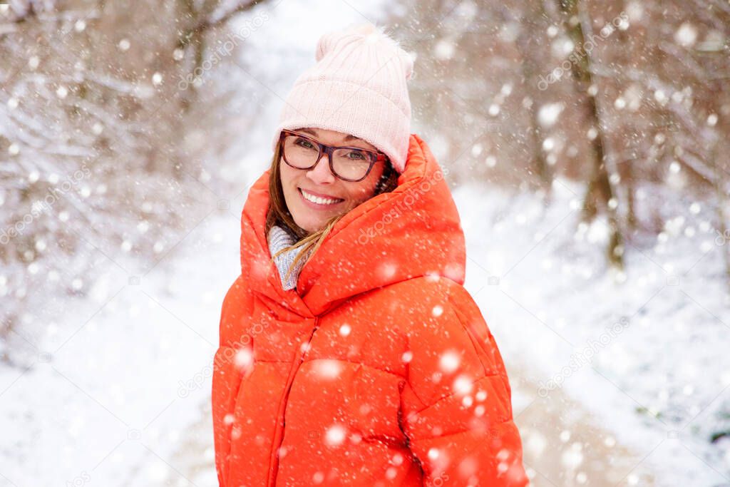 Close-up shot of a happy middle aged woman wearing hat while standing outdoor and enjoy snowfall. 