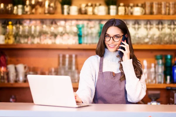 一个漂亮的女服务员站在餐馆柜台后面用手机和笔记本电脑刻画的形象 小商业 — 图库照片