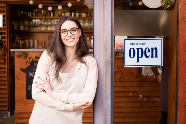 笑顔の若いカフェショーのオーナーの女性の腕を戸口に横切って立ってのショット ガラスの扉にサインを — ストック写真