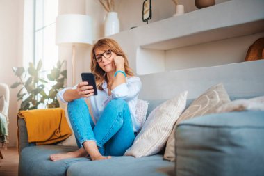 Düşünen bir kadının evde koltukta dinlenirken cep telefonunu ve mesajlarını kullanması..