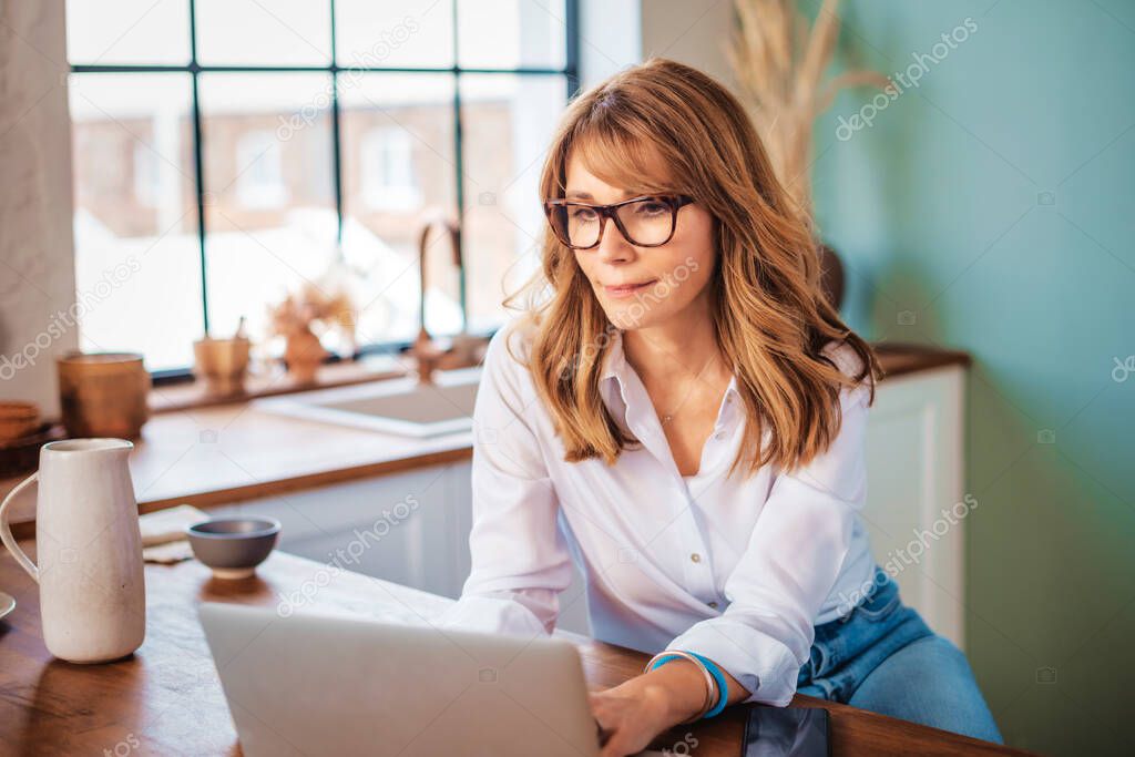 An attractive middle aged woman using her laptop while relaxing at home in her kitchen. 