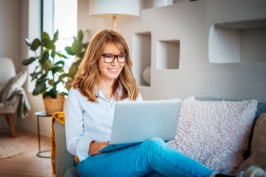 Orta yaşlı bir kadının evdeki kanepede dizüstü bilgisayar kullanması. İş kadını evden çalışıyor..