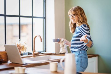 Orta yaşlı bir kadının laptopunu kullanırken ve mutfakta kahvaltı hazırlarken görüntülü konuşma yaparken..