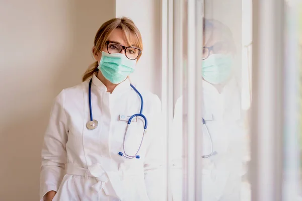 Female doctor wearing face mask and looking careworn while standing at the window.