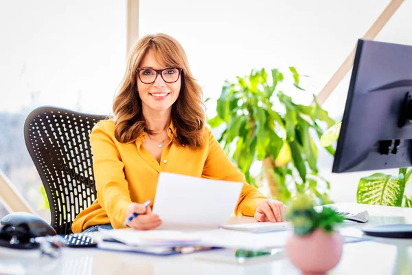 幸せな成熟したビジネスマンのショットオフィスの机に座って オフィスでコンピュータ上で作業しながら いくつかの書類を行う女性 — ストック写真