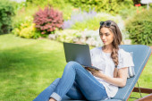 Lövés fiatal nő ül a napozóágyon a kertben, és használja a laptopját. 