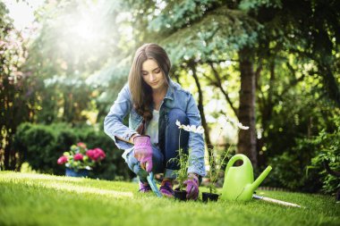 Arka bahçede bahçeyle uğraşan genç bir kadının tam boy çekimi. Çiçek dikerken gündelik kıyafetler giyen mutlu kadın..