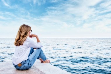 Arka planda, deniz kenarında oturmuş denize bakan sıradan kıyafetler giyen bir kadının fotoğrafı var..
