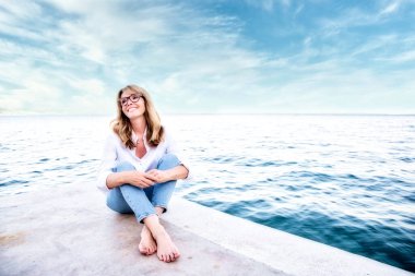 Tam boy, orta yaşlı, beyaz gömlek ve kot pantolon giyen, deniz kenarında dinlenen bir kadın..