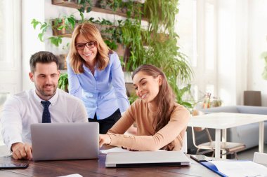 Bir grup iş adamı iş projesinde birlikte çalışıyor. Genç sekreter kadın ve finans danışmanı iş adamı dizüstü bilgisayarın önünde otururken orta yaşlı profesyonel kadın arkalarında duruyor ve tavsiyelerde bulunuyor..