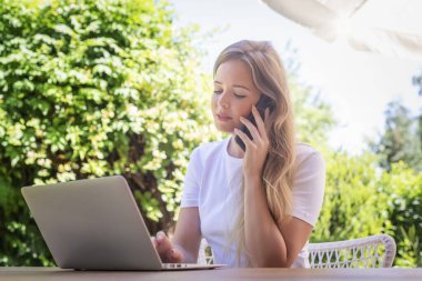 Evde arka bahçede oturup çalışırken cep telefonu ve defter kullanan çekici genç bir kadın. Merkez Ofis.