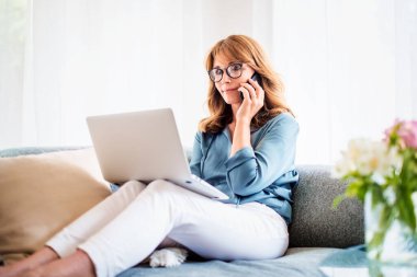 Orta yaşlı iş kadını dizüstü bilgisayar kullanıyor ve evde bir koltukta oturup çalışırken iş görüşmesi yapıyor. Merkez Ofis.