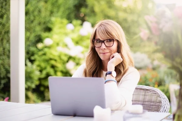 快乐而成熟的女人坐在花园的家中 使用笔记本电脑 总部办公室 — 图库照片