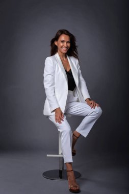 Stüdyoda, beyaz süit giyen mutlu ve çekici bir kadının koyu gri arka planda oturduğu uzun bir çekim.. 