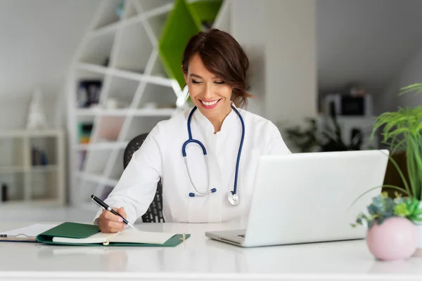 Lächelnde Gesundheitshelferin Die Papierkram Erledigt Und Laptop Benutzt Während Sie — Stockfoto