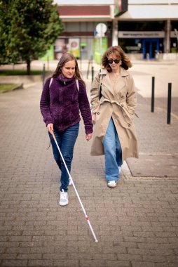 Görme engelli ve işitme engelli bir kadın, kız arkadaşıyla şehirde yürüyor. Görme engelli bir kadın elinde beyaz bir baston tutuyor ve konuşuyor. Tam boy çekim. 