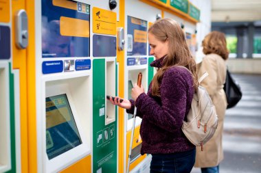 Tren istasyonunda bilet makinesinin önünde durup tren bileti alan görme engelli bir kadın. Kendine güvenen bir kadın işitme cihazı takıyor ve elinde beyaz bir baston tutuyor. Cep telefonuyla ödeme yapmak. Temassız ödeme. 