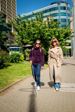 Görme engelli ve işitme engelli bir kadın, kız arkadaşıyla şehirde yürüyor. Görme engelli bir kadın elinde beyaz bir baston tutuyor ve konuşuyor. Tam uzunluk. 