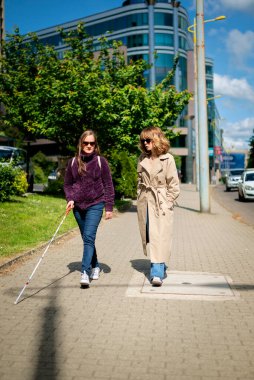Görme engelli ve işitme engelli bir kadın, kız arkadaşıyla şehirde yürüyor. Görme engelli bir kadın elinde beyaz bir baston tutuyor ve konuşuyor. Tam uzunluk. 