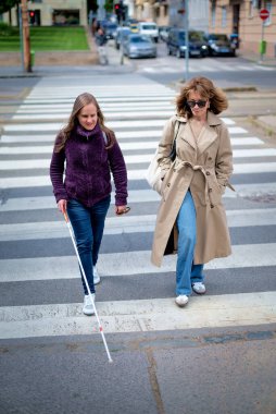 Görme engelli bir kadın yaya geçidinde kadın arkadaşıyla yürüyor. Görme engelli arkadaşına yardım eden olgun, beyaz bir kadın. Tam uzunluk. 