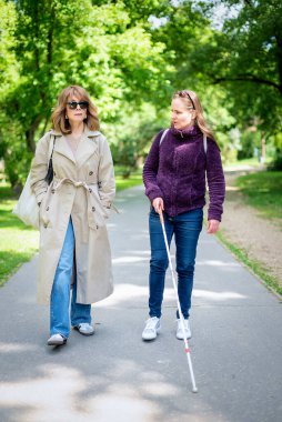 Şehir parkında kadın arkadaşıyla yürüyen görme engelli bir kadın. Görme engelli arkadaşına yardım eden olgun, beyaz bir kadın. Tam uzunluk. 