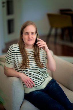 Akıllı telefonlu görme engelli bir kadın rahat evindeki koltukta oturuyor. Gülümseyen kadın günlük kıyafetler giyiyor..