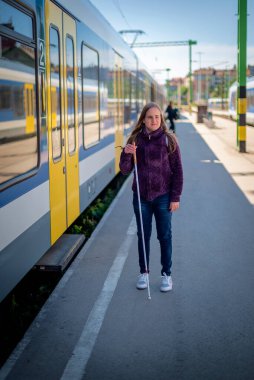 Tren istasyonunda demiryolu platformunda yürüyen beyaz bastonlu kör kadın portresi. Treni bekleyen görme engelli bir kadın. Günlük kıyafetler. Tam boy çekim. 