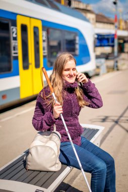 Beyaz bastonlu kör kadın portresi. Şehrin dışındaki tren istasyonunda bankta oturuyor. Treni bekleyen ve telefon kullanan görme engelli bir kadın. Günlük kıyafetler.. 