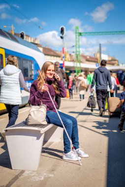 Beyaz bastonlu kör kadın portresi. Şehrin dışındaki tren istasyonunda bankta oturuyor. Treni bekleyen ve telefon kullanan görme engelli bir kadın. Günlük kıyafetler. Tam uzunluk. 