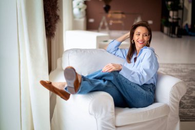 Açık mavi gömlek ve kot pantolon giyen kendinden emin ve çekici orta yaşlı bir kadın. Kendine güvenen bir kadın modern evinde rahat bir koltukta dinleniyor.. 