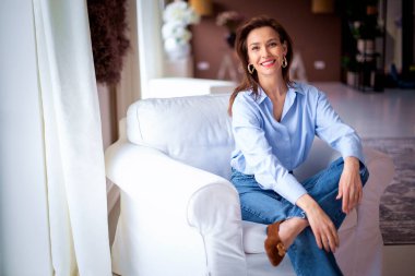 Açık mavi gömlek ve kot pantolon giyen kendinden emin ve çekici orta yaşlı bir kadın. Kendine güvenen bir kadın modern evinde rahat bir koltukta dinleniyor.. 