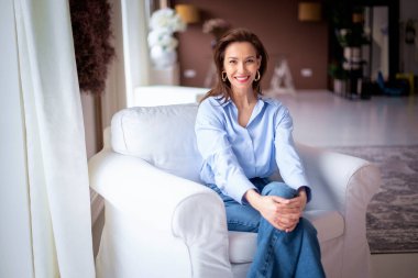 Açık mavi gömlek ve kot pantolon giyen kendinden emin ve çekici orta yaşlı bir kadın. Kendine güvenen bir kadın modern evinde rahat bir koltukta dinleniyor.. 