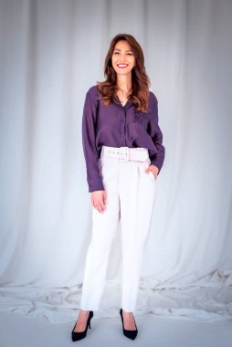 Gülümseyen olgun bir kadının portresi. Esmer saçlı, kendine güveni tam bir kadın. Gömlek ve beyaz pantolon giyen çekici bir kadın. Uzayı kopyala Tam boy çekim.