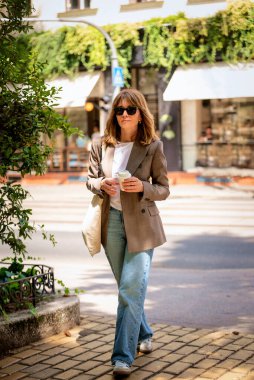 Sıradan giysiler içinde orta yaşlı stil sahibi bir kadın, bir fırının önünde şehir caddesinde yürüyor. Kendine güvenen bir kadın elinde bir kahve fincanı tutuyor ve kameraya bakıyor. Tam uzunluk.