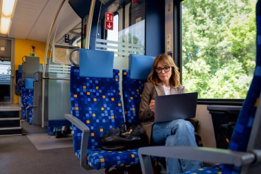 Dizüstü bilgisayarında çalışan sarışın bir kadın ve trenle seyahat ederken akıllı telefon kullanıyor. Modern bir trende tek başına oturan kendinden emin bir kadın. Uzak çalışma.